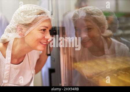 Die junge Frau als Bäckerlehrling in der Ausbildung prüft Backwaren im Ofen Stockfoto