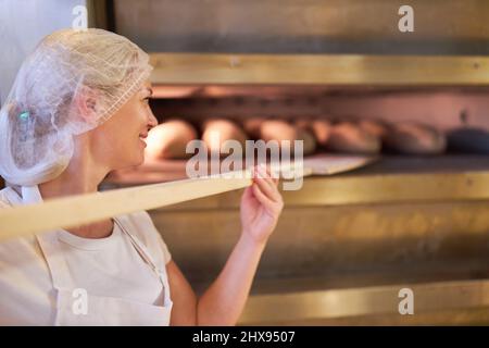 Die junge Frau als Bäckerlehrling in der Ausbildung schiebt in der Bäckerei Brot in den Ofen Stockfoto