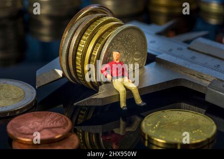 Miniaturmenschen: Junger Mann, der auf einem Messschieber sitzt und den Münzstapel misst. Altersvorsorge. Geld sparen und investieren. Die Zeit wird für heruntergezählt Stockfoto