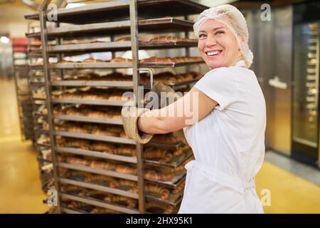 Eine fröhliche junge Frau schiebt als Bäckerlehrling fertig gebackene Croissants in die Bäckerei Stockfoto