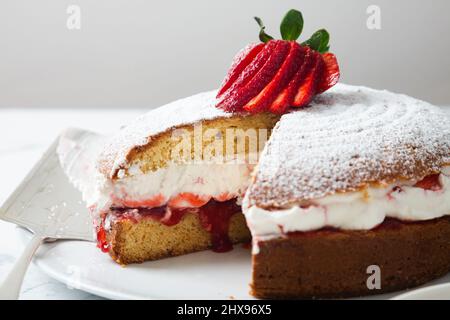 Hausgemachter Viktoria-Biskuitkuchen, gefüllt mit Erdbeeren, Marmelade und Schlagsahne, verziert mit Puderzucker und Erdbeere auf einem weißen Marmortisch Stockfoto