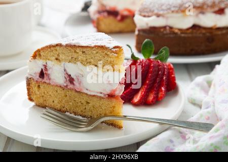 Eine Portion Victoria-Biskuitkuchen, gefüllt mit Erdbeeren, Marmelade und Schlagsahne, verziert mit Puderzucker und Erdbeere auf einem weißen Holztisch Stockfoto