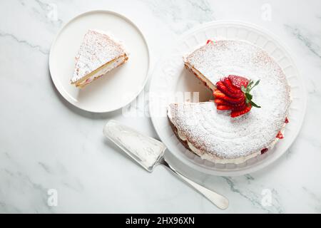 Blick von oben auf den hausgemachten Victoria-Biskuitkuchen, gefüllt mit Erdbeeren, Marmelade und Schlagsahne, verziert mit Puderzucker und Erdbeere auf weißem Marmor Stockfoto