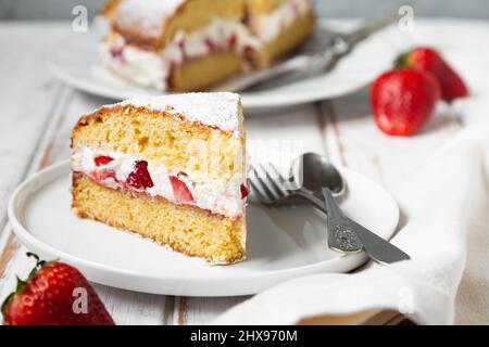 Hausgemachter Viktoria-Biskuitkuchen, gefüllt mit Erdbeeren, Marmelade und Schlagsahne, verziert mit Puderzucker und Erdbeere auf einem weißen Holztisch Stockfoto