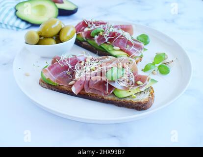 Vollkornbrot-Sandwiches mit Scheiben Avocado, Prosciutto oder Jamon, gekeimten Samen und Kräutern. Sauberes Essen, gesundes Frühstück. Draufsicht. Stockfoto