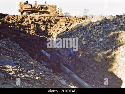 Zwei Männer auf einer Baustelle, die mit einem großen Rohr arbeiten, ein Bulldozer im Hintergrund ca. 1960 Stockfoto