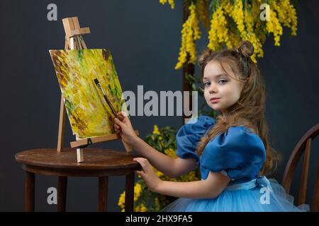 Ein Mädchen sitzt in der Nähe einer Mimose und malt ein Bild in einem blauen Kleid Mimosa Pinsel Mädchen, aus Kleid hält in Schönheit für Lächeln glücklich, blonde Blüte. 8 Stockfoto