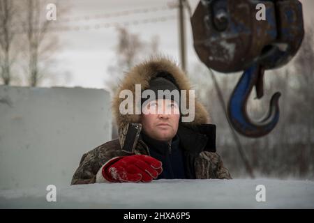 Porträt eines Arbeiters in einer Winterjacke mit einer Kapuze am Haken eines LKW-Krans beim Bau einer Eisstadt Stockfoto