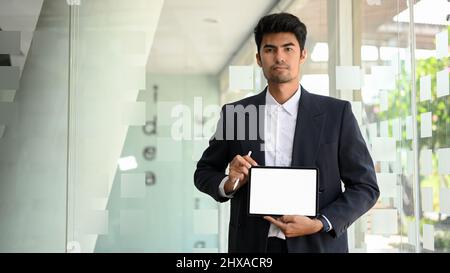 Asiatischer Geschäftsmann oder leitender Manager in formellem Anzug, der vor dem Besprechungsraum steht und eine Hand zeigt, die ein Modell des Tablet-Bildschirms zeigt. Stockfoto