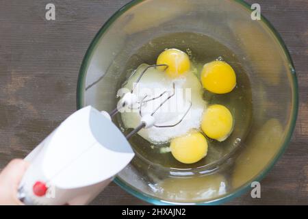 Frau hält den Mixer in der Glasschüssel mit Zucker und Eiern, bevor sie schlägt Stockfoto