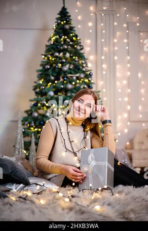 Eine junge Frau in einem Pullover mit langen, schönen Haaren sitzt und lacht vor Weihnachtsgeschenken. Frohes Neues Jahr Stockfoto