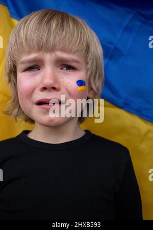 Weinender, verängstigter Junge vor dem Hintergrund einer gelb-blauen ukrainischen Flagge. Russlands Invasion der Ukraine, ein Plädoyer für Hilfe für die Weltgemeinschaft. c Stockfoto