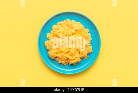 Rühreier Teller minimalistisch auf einem gelben Tisch, Blick von oben. Köstliche hausgemachte Rühreier auf einem blauen Teller isoliert auf einem gelben Hintergrund. Stockfoto
