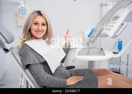 Besuch in der Zahnklinik. Junge positive weibliche Klientin zeigt Daumen nach oben und lächelt während der zahnärztlichen Untersuchung und Konsultation in der Zahnarztpraxis. Kopie Stockfoto