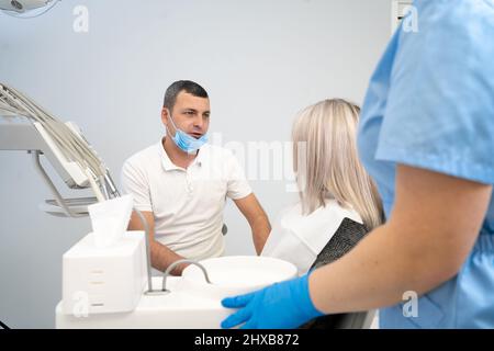 Schönes kaukasisches Mädchen mit Zahnschmerzen sitzt in einem Zahnarztstuhl. Ein professioneller männlicher Zahnarzt hilft seiner Patientin bei Schmerzen. Schmerzhafte Hilfe bei der Behandlung Stockfoto