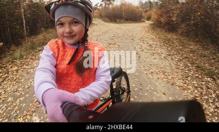 Machen Sie Selfie auf dem Kameratelefon. Ein kaukasisches Kind fährt im Herbstpark auf der Fahrradstraße. Mädchen reiten schwarz orange Zyklus im Wald. Kind geht Fahrrad spor Stockfoto