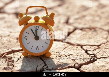 Wecker mit fünf Minuten vor zwölf Uhr auf einem geknackten, öden Boden. Konzept des Klimawandels oder der globalen Erwärmung. Stockfoto