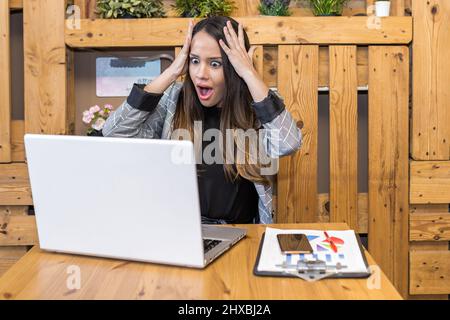 Erstaunte junge Freiberuflerin mit Kopf und Öffnung des Mundes beim Lesen überraschender Daten auf dem Netbook-Bildschirm während der Fernarbeit im Café Stockfoto