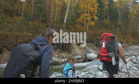 Die Menschen gehen in der Nähe des Bergflusses. Familienreisen. Menschen Umwelt durch Berge, Flüsse, Bäche. Eltern und Kinder laufen mit Wanderstöcken. Mann Stockfoto
