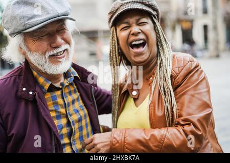 Multirassische ältere Paar mit Spaß im Freien mit Stadt im Hintergrund - Liebe, Beziehung und fröhliche ältere Lifestyle-Konzept - Fokus auf japanischen Mann Stockfoto