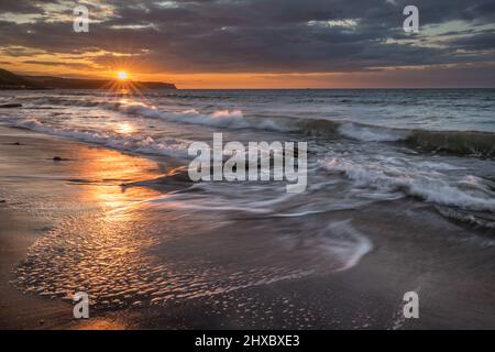 Golden Hour Sonnenuntergang am Whitby Beach. Warmer Himmel und sanfte, sanfte Wellen entlang einer sandigen Küste mit großen Felsen. Stockfoto