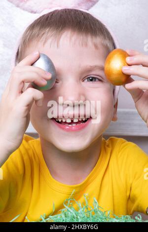 Porträt eines kleinen blonden Jungen, der seine Augen mit bunten Eiern bedeckt, Nahaufnahme, vertikaler Rahmen. Osterurlaub mit der Familie. Das Kind hat Spaß und feiert Stockfoto