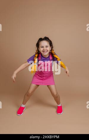 Überschwängliches kleines weibliches Kind mit gelben Kanekalon-Zöpfen, die Beine weit auseinander und lächelnd zeigen, dass es Piece-Zeichen mit Händen in rosa Jumpsuit trägt Stockfoto