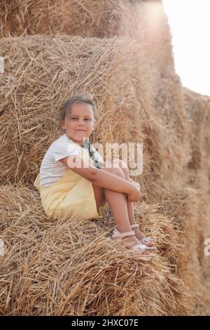 Porträt eines friedlichen hübschen jungen Mädchens auf Heuhaufen mit einem Lächeln und blinden Augen, die auf die Kamera schauen und Heu in den Händen halten, in der Sundress tragend. Spaß haben Stockfoto