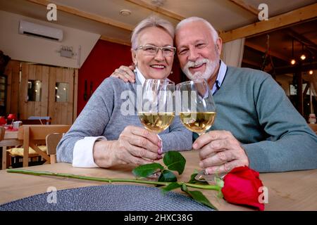 Ein reifes, romantisches Paar kaukasischer Männer und Frauen sitzt in einem Restaurant. Verliebte Senioren, die die Kamera betrachten, Gläser mit Wein toasten und anklirren. Stockfoto