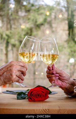Die Hände eines älteren romantischen Paares, die Gläser mit Wein halten, toasten und mit ihnen über einer roten Rose klirren. Hochzeitstoast. Stockfoto