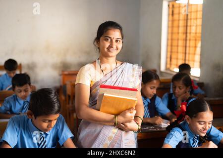 Lächelnder Lehrer mit Büchern in der Hand, die im Klassenzimmer zwischen den Schülern im Klassenzimmer stehen - Konzept des beruflichen Berufs, der Ermächtigung der Frau und Stockfoto