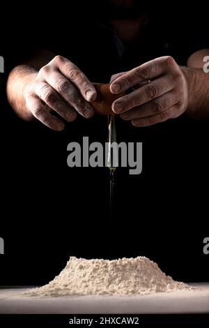 Ein Mann zerbricht ein Ei in Mehl, um vor dunklem Hintergrund Teig auf einem Tisch zu machen. Stockfoto