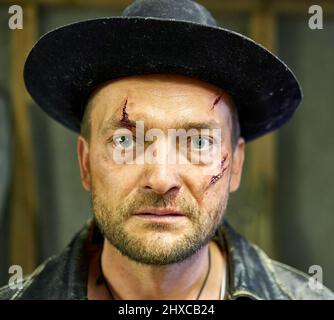 CIEMUPE, LETTLAND - 11. JULI 2020: Ein Mann mit blutigen Wunden im Gesicht im alten schwarzen Hut und in der Jacke während der Fotosession Stockfoto