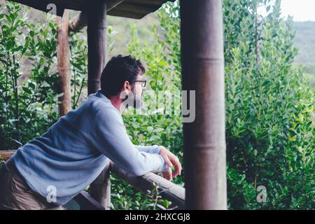 Ein Mann steht zu Hause auf der Terrasse und bewundert die grünen Bäume und den natürlichen Wald. Stockfoto