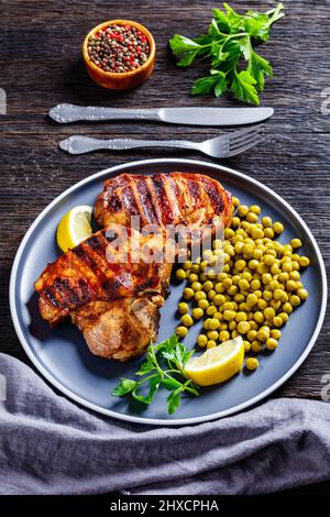 Gegrillte Schweinefleisch-T-Bone-Steaks mit gekochten grünen Erbsen und Zitrone auf dem Teller auf einem Holztisch, vertikale Ansicht von oben, Nahaufnahme Stockfoto