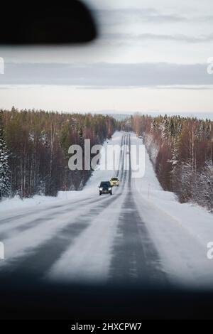 Autos fahren geradeaus auf schneebedeckter Straße mit Wald auf der linken und rechten Seite in Finnland Stockfoto