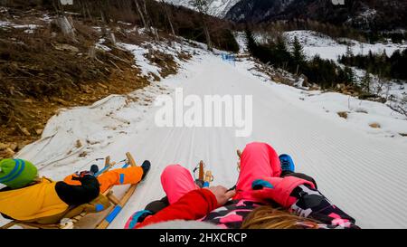 Eltern, die ein Kind auf einem Schlitten fahren, sich schnell auf der holprigen Schneestrecke bewegen, aus der Perspektive aufgenommen. Stockfoto