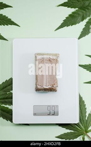 Abgepackte Haschisch-Tablette auf Waage, Marihuana-Blätter, grüner Hintergrund Stockfoto