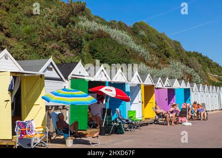 England, Dorset, Bournemouth, Bournemouth Beach, Menschen, die vor einer Reihe von farbenfrohen Strandhütten sonnenbaden Stockfoto