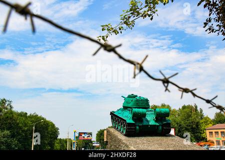 Denkmal für den alten sowjetischen Panzer T-34. Ein Militärpanzer steht auf einem Sockel vor dem Hintergrund eines blauen Himmels, Stacheldrahts, grüner Bäume und einer Stadtstraße. Stockfoto