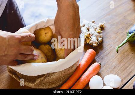 Nahaufnahme eines Mannes, der in der Küche seines Hauses steht und Kartoffeln auswählt, die sich in einem Sack befinden, zusammen mit anderem Gemüse auf dem Tisch Stockfoto
