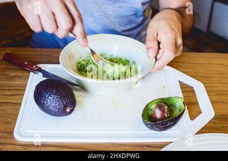 Nahaufnahme kaukasischer Mann, der in seiner Küche sitzt und Guacamole mit einer Gabel in einer weißen Schüssel zubereitet Stockfoto