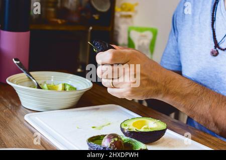 Nahaufnahme kaukasischer Mann, der in seiner Küche sitzt und Avocado hackt, um Guacamole für das Mittagessen zuzubereiten. Stockfoto