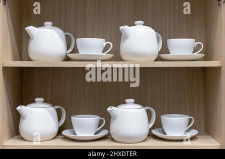 Weiße Keramikbecher mit Untertassen und Teekannen auf einem Regal in einem Holzschrank. Stockfoto