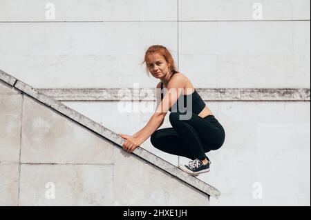 Parkour Frau klettert das Gebäude hinauf und schaut in die Kamera. Frau beim Parkour in der Stadt an einem sonnigen Tag. Vertikales Foto Stockfoto