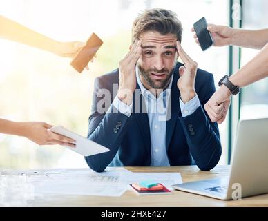 Zu viel zu tun, zu wenig Zeit. Porträt eines jungen Geschäftsmannes, der an einem Schreibtisch sitzt, umgeben von Händen, die Gegenstände halten. Stockfoto