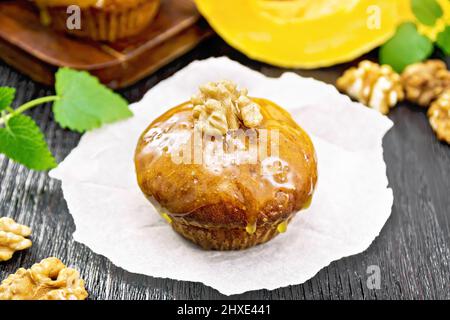 Kleiner Kürbis-Cupcake mit orangefarbener Glasur und Walnüssen auf Pergament, Minze-Zweige auf dunklem Holzbrett-Hintergrund Stockfoto