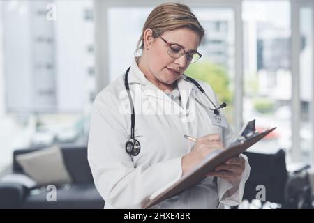 Ihren Zeitplan zu sortieren. Ausgeschnittene Aufnahme einer attraktiven jungen Ärztin, die in ihrem Büro auf einem Klemmbrett schreibt. Stockfoto