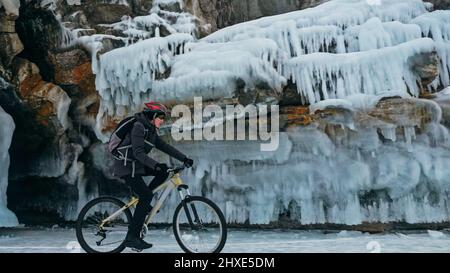 Der Mensch fährt Fahrrad in der Nähe der Eisgrotte. Felsen mit Eishöhlen Eiszapfen. Teenage ist in schwarzer Daunenjacke, Fahrradrucksack, Helm gekleidet. Reifen auf covere Stockfoto