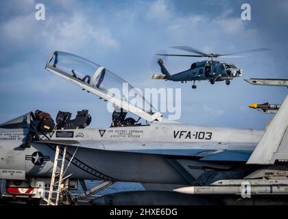 220306-N-UD000-1077 ATLANTISCHER OZEAN (6. März 2022) Ein Segler, der mit dem Strike Fighter Squadron (VFA) 103 beauftragt wurde, wischt das Cockpit eines F/A-18 F Super Hornet-Flugzeugs an Bord des Flugzeugträgers USS George H.W. Bush (CVN 77), während ein MH-60R Sea Hawk Hubschrauber, der dem Hubschrauber Maritime Strike Squadron (HSM) 48 zugewiesen wurde, im Hintergrund fliegt, März 6. George H.W. Bush stellt der nationalen Befehlsbehörde flexible, anpassungsfähige Kampfkapazitäten für den Krieg durch die Trägerstreikgruppe zur Verfügung, die die maritime Stabilität und Sicherheit aufrechterhält, um den Zugang zu gewährleisten, Aggressionen abzuschrecken und die Vereinigten Staaten zu verteidigen Stockfoto